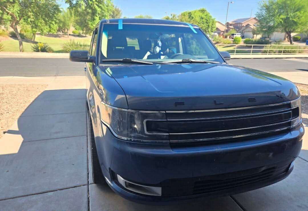 Dark blue Ford Flex with a freshly installed windshield secured with blue tape, sunny Buckeye driveway.