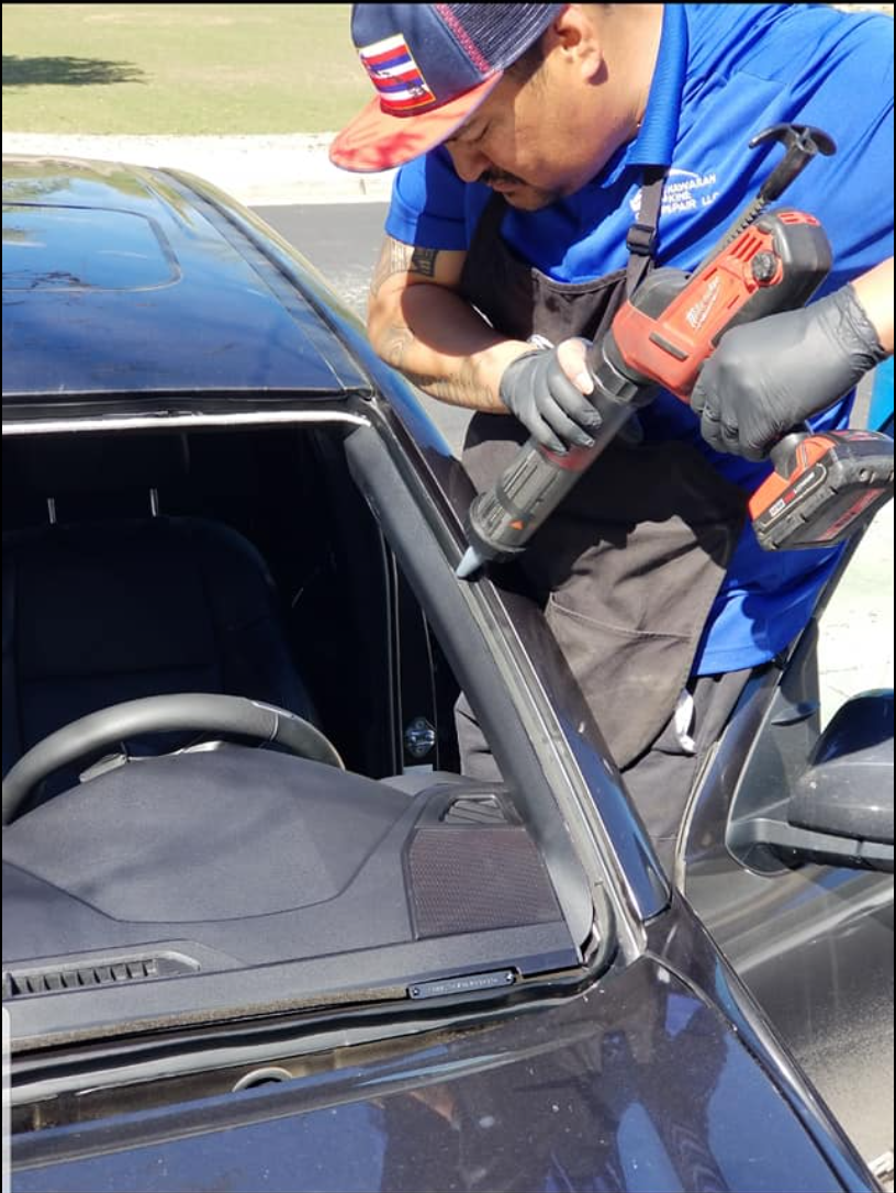 Henry working from inside a vehicle with a drill, removing trim during a windshield job.